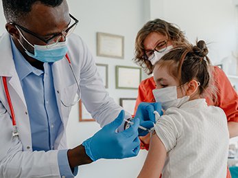 A child in a white shirt getting a vaccine from a doctor with blue gloves, a white lab coat and a red stethoscope. The guardian of the child watches attentively and with curiosity.
