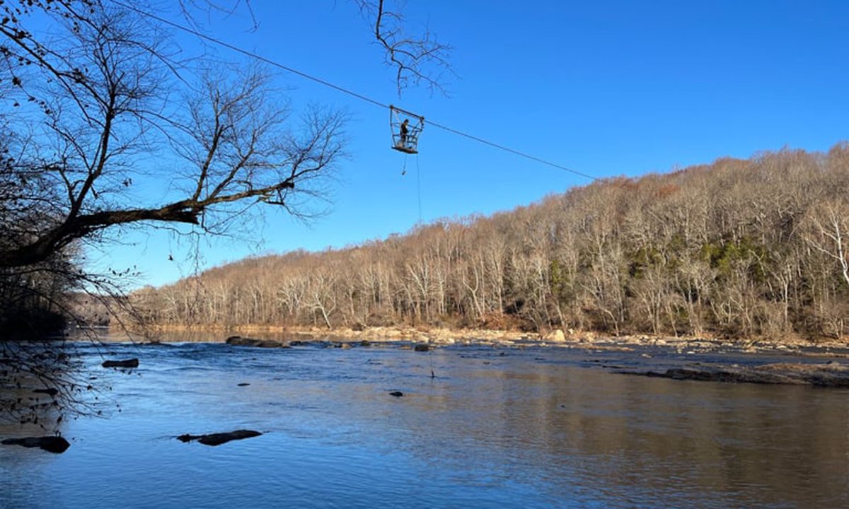 A far-away photo of a man collecting samples from a wide river while suspended above it by a wire.