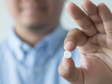Man holding small, round white pill up to the camera in between his pointer finger and thumb. His face is partially visible but blurry in the background.