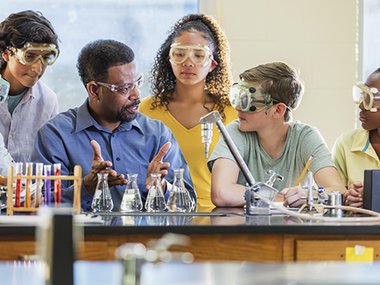 Lab teacher talking to students gathered around lab table in a lab class. Different lab equipment is on top of the table such as test tubes and other glass equipment.