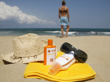 An image showing a bottle of sunscreen, sunglasses and hat resting on a folded towel. The items are sitting in the sand. A blurry outline of a person in the background is walking toward water.