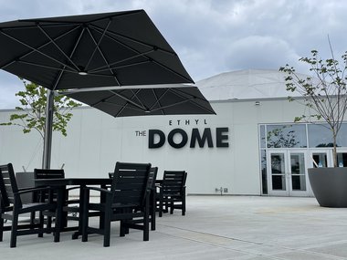 An image of the Science Museum's Dome Plaza. A black recycled plastic table with a dark umbrella shading it is to the left. The words "The Dome" are visible on the wall in the background. The concrete is bright white.