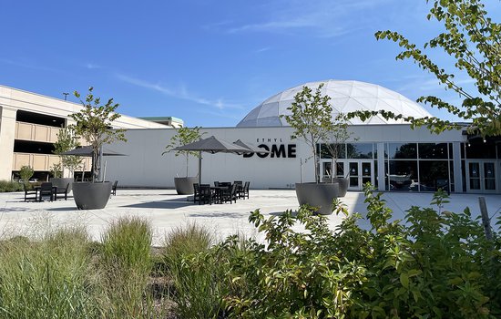 An image of the Science Museum's Dome Plaza showing black recycled plastic tables with a dark umbrellas shading them. The words "The Dome" are visible on the wall in the background. The concrete is bright white.