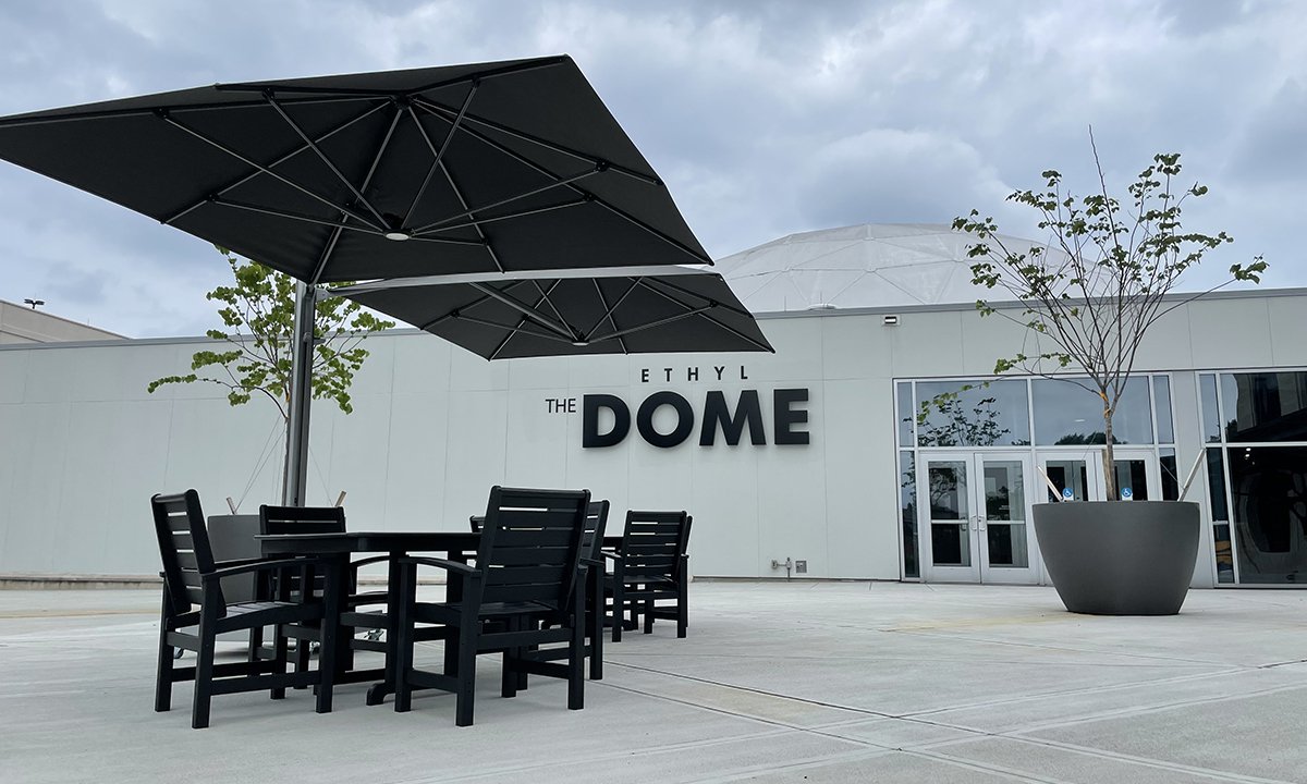 An image of the Science Museum's Dome Plaza showing black recycled plastic tables with a dark umbrellas shading them. The words "The Dome" are visible on the wall in the background. The concrete is bright white.