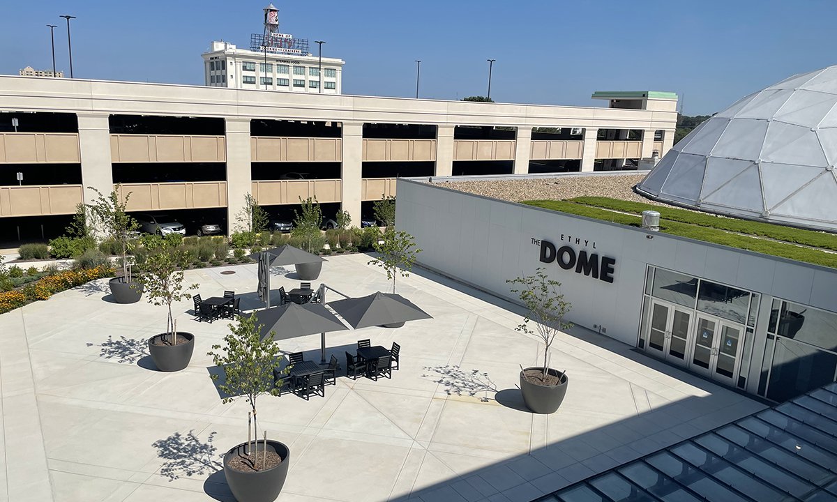 An image of the Science Museum's Dome Plaza showing black recycled plastic tables with a dark umbrellas shading them. The words "The Dome" are visible on the wall in the background. The concrete is bright white.