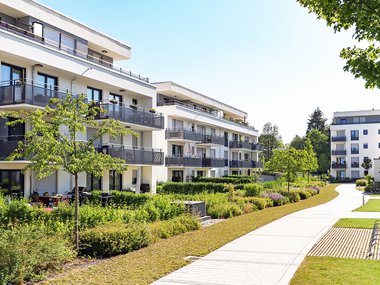 Photograph showing an apartment building to the left and a walking path to the right. A blue sky is above. A tree is poking out of the top right corner. The building and sidewalk are white to reflect sunlight. There is plentiful green landscaping.