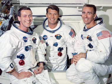 A photograph of the Apollo 7 crew" three men in their white space suits sitting in the spacecraft looking at the camera.