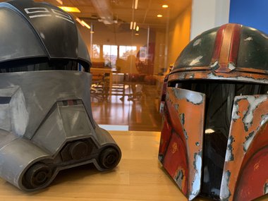 Two Star Wars cosplay helmets sitting on a table. The right one is orange, the left one is dark green.