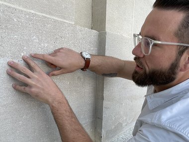 A photograph of a man with dark hair, glasses and a white shirt looking at a wall and pointing to something not immediately visible.
