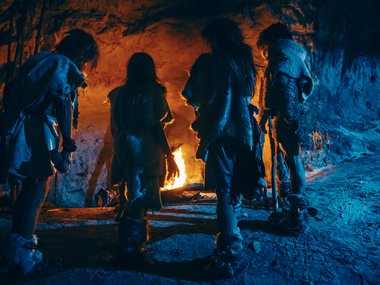 An illustration of early humans standing in the mouth of a cave looking at a burning fire. Their backs are to the viewer.