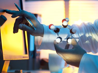 Image of hands holding a shape with light beams streaming in. A computer is in the background. It looks like a lab.