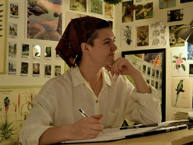 Scientific Illustrator Nick Garnhart working in their studio. Her profile is to the camera. Her walls are covered in prints. They are wearing a white shirt and have a head covering.