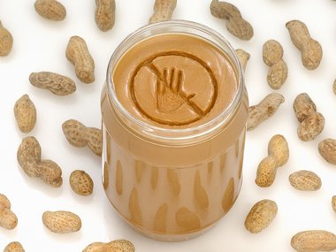 A peanut butter jar with the lid off. A caution symbol is carved into the peanut butter. Whole peanuts are spread on the ground around the outside of the jar.