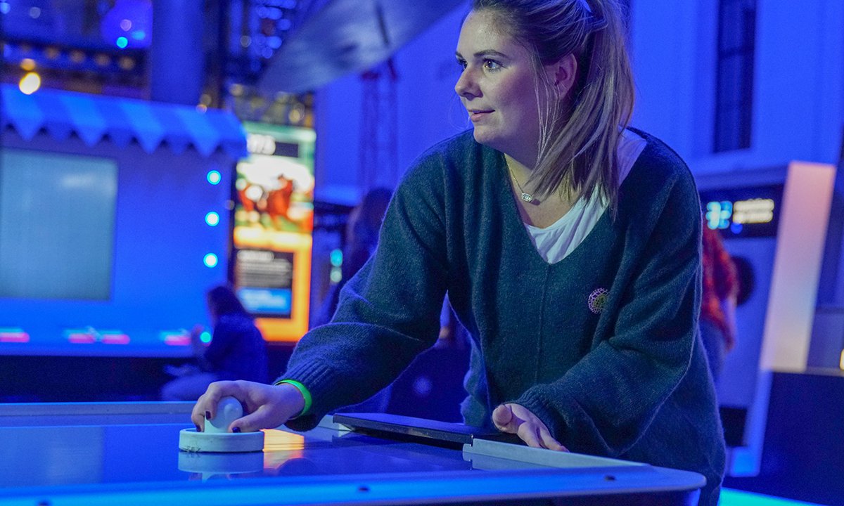 A guest with blonde hair in a ponytail and green sweater plays air hockey against a robot in the Speed exhibit in the science museum.