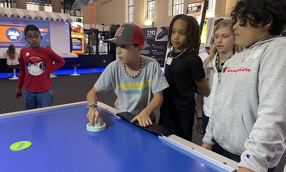 One student hits the puck on the air hockey table as four students watch (three to the right and one to the left).