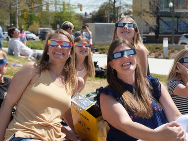 Guests sitting on the Science Museum front lawn wearing eclipse viewing glasses smiling at the camera.