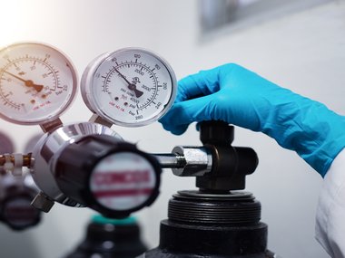 person wearing blue gloves and a white lab coat adjusting a gas regulator on top of a cylinder with two pressure gauges visible