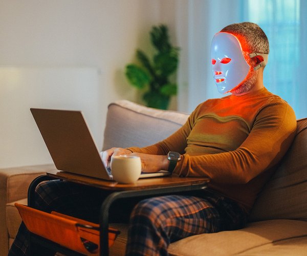 Person sitting on a couch wearing a red light therapy mask while using a laptop that sits on their lap