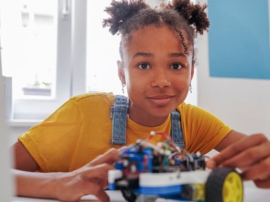 A young girl with braided pigtails, wearing a golden yellow t-shirt and denim overalls, grins at the camera asthey build or assemble a small robot with wheels