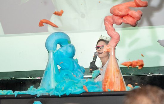 A woman wearing a crown and pink googles watching blue and red chemical reactions.