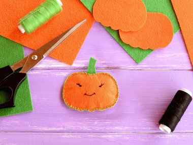 Photograph of scissors and orange felt on a purple background with a orange pumpkin plushie in the center