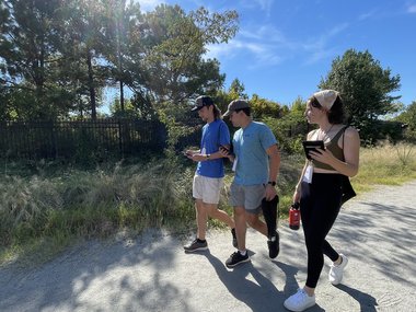 Three University of Richmond students walking on the Science Museum's ProtoPath collecting air quality data using a white sensor and black tablet.