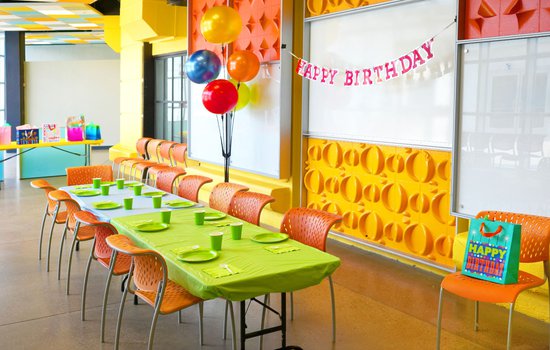 Room with large bright windows and orange green and yellow colored chairs and wall decorations featuring birthday gifts in the Challenge Lab