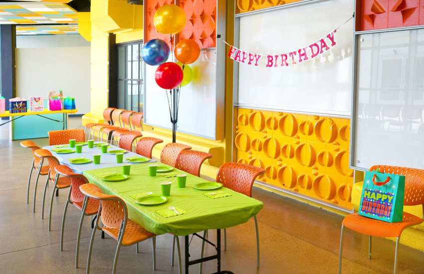 Room with large bright windows and orange green and yellow colored chairs and wall decorations featuring birthday gifts in the Challenge Lab
