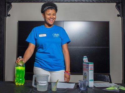 A female volunteer standing behind a table that has ingredients and containers. There is a box of Borax to the right. The volunteer's hand rests on the top of a 2-liter plastic bottle full of bright green liquid. The female is wearing a blue shirt.