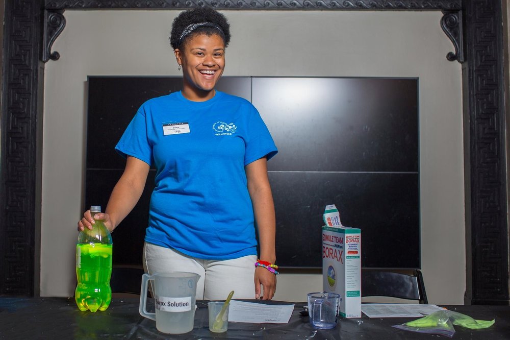 A female volunteer standing behind a table that has ingredients and containers. There is a box of Borax to the right. The volunteer's hand rests on the top of a 2-liter plastic bottle full of bright green liquid. The female is wearing a blue shirt.