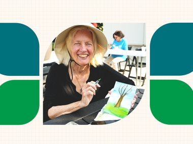 older woman, wearing a sun hat, smiles while showing off her artwork of a hand painted tree