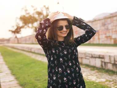 A woman wearing sunglasses and a long sleeve floral black dress places her hands on top of the white hat on her head