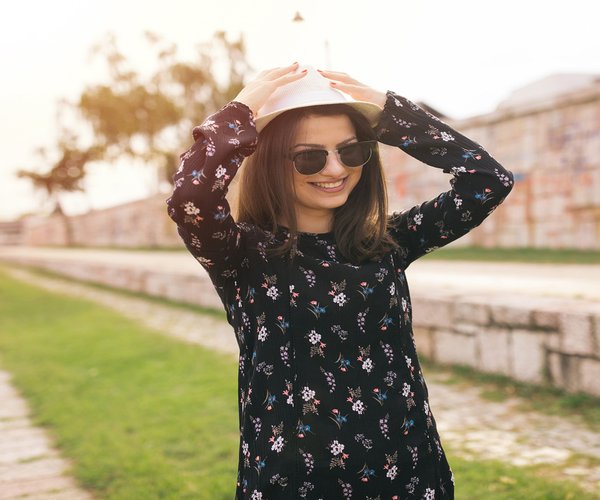 A woman wearing sunglasses and a long sleeve floral black dress places her hands on top of the white hat on her head