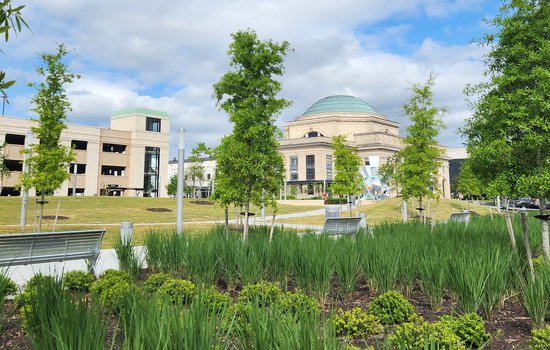 A color photograph of The Green, the Science Museum of Virginia's community greenspace. Trees, grass, shrubs and flowers are visible, with the building in the background and a blue sky.