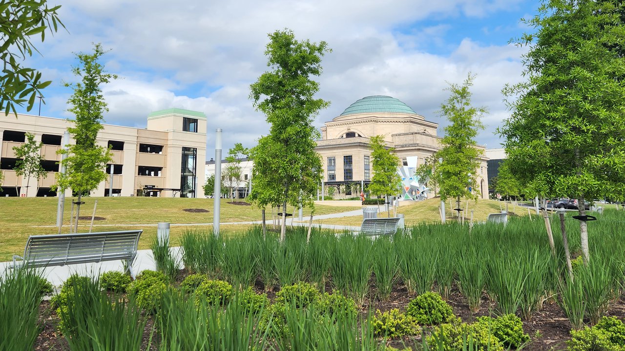 A color photograph of The Green, the Science Museum of Virginia's community greenspace. Trees, grass, shrubs and flowers are visible, with the building in the background and a blue sky.