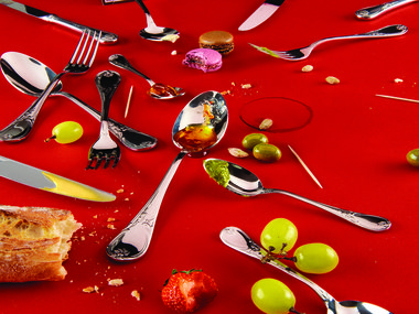 Spoons, knives and forks laid out across a red background. Dispersed throughout are olives, macaroons, bread and grapes