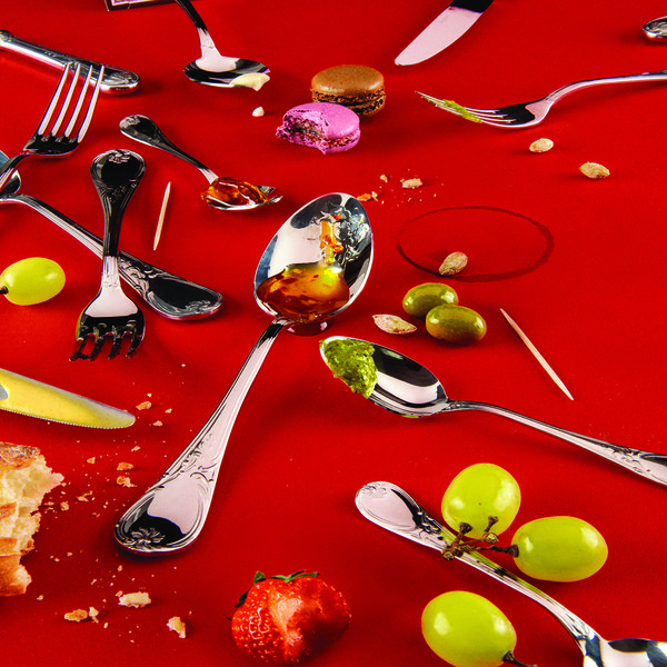 Spoons, knives and forks laid out across a red background. Dispersed throughout are olives, macaroons, bread and grapes