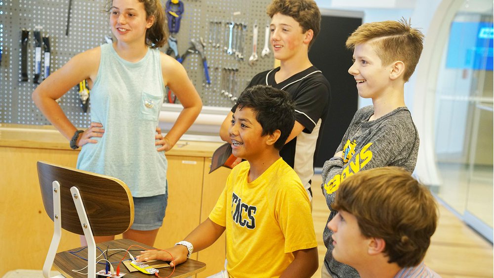 Five children standing in front of a wall with tools hanging on it, smiling excitedly.