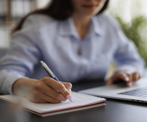 woman wearing a collared shirt writes in a notebook while also using the trackpad on a laptop