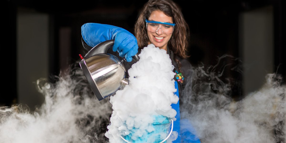 Woman with googles and blue safety gloves pouring water from a silver kettle into a blue tub of liquid nitrogen. It has created a large plume of steam that has spread throughout the space.
