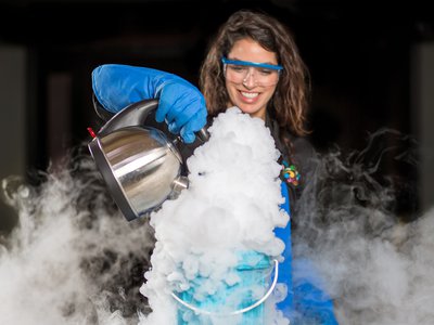 Woman with googles and blue safety gloves pouring water from a silver kettle into a blue tub of liquid nitrogen. It has created a large plume of steam that has spread throughout the space.