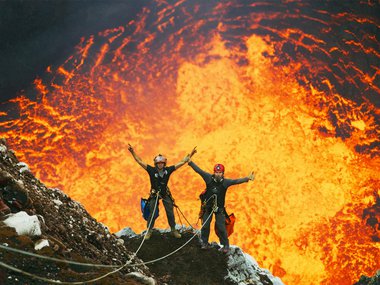 Two people in front of a volcano and exploding magma.