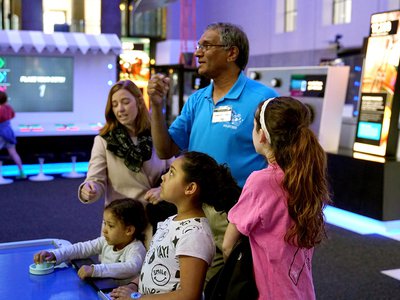 A male volunteer wearing a blue shirt is standing in the middle of four people: an adult woman to the left, two smaller children in the middle, and a teen girl to the right. They are all looking at the air hockey robot in the Speed exhibition.