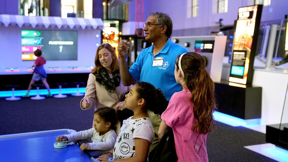 A male volunteer wearing a blue shirt is standing in the middle of four people: an adult woman to the left, two smaller children in the middle, and a teen girl to the right. They are all looking at the air hockey robot in the Speed exhibition.