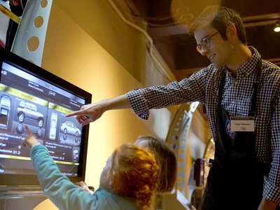 Male volunteer pointing to a screen to show a young guest how to use the interactive unit in an exhibition.
