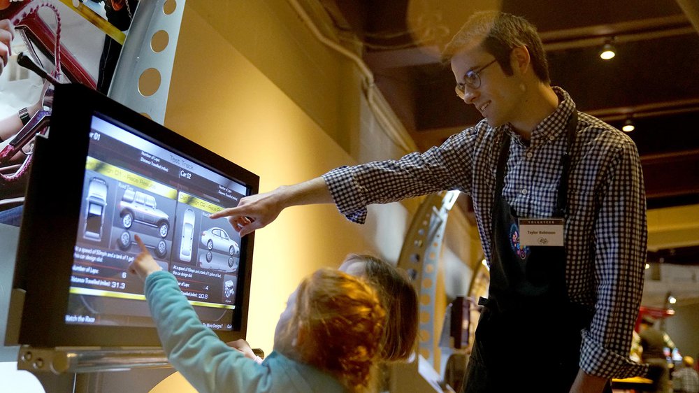 Male volunteer pointing to a screen to show a young guest how to use the interactive unit in an exhibition.
