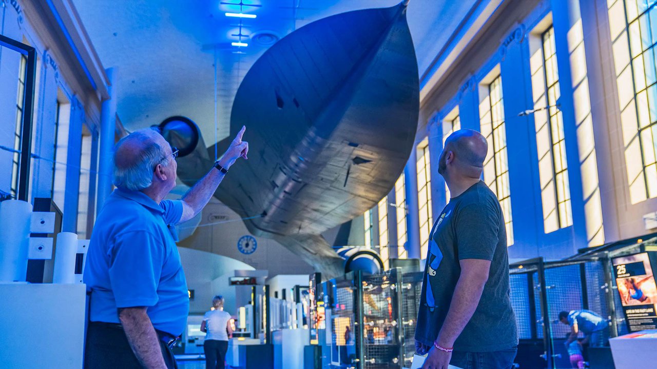 A volunteer is standing to the right of the SR-71 pointing at to to the guest standing on the right. They are both in the Speed exhibition. Their side is to the camera and they are looking away and up at the jet hanging from the ceiling.